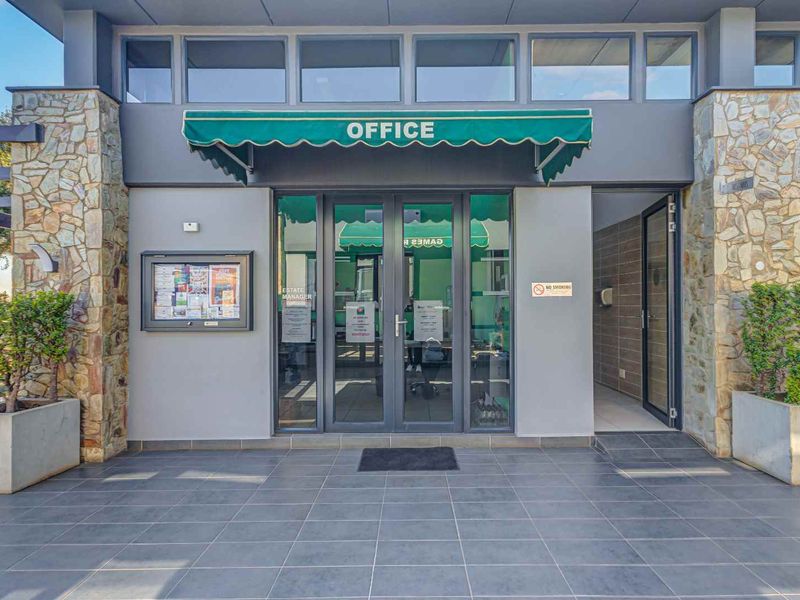 The entrance to the estate office featuring stone-clad pillars, a green awning, and modern glass double doors with a tiled patio area.