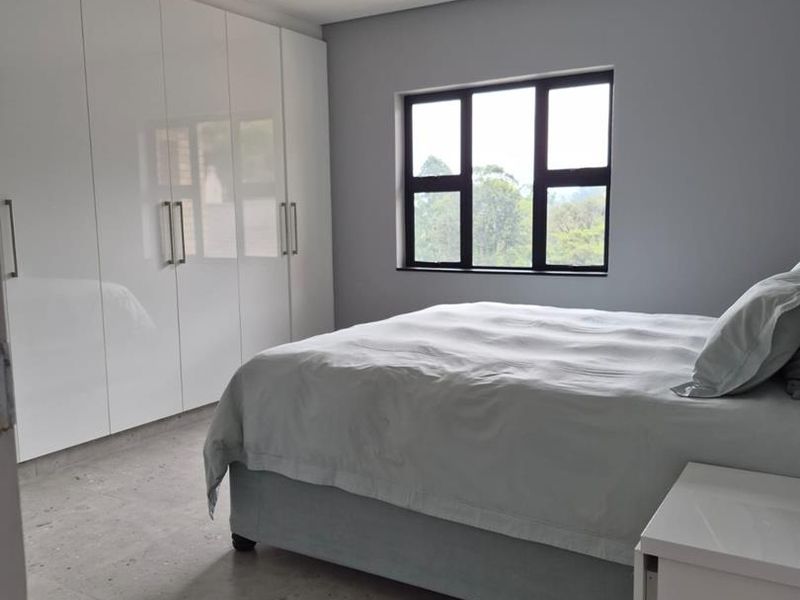 A modern bedroom featuring sleek white built-in cupboards, neutral tiled flooring, and a large window providing natural light and a view of the greenery.
