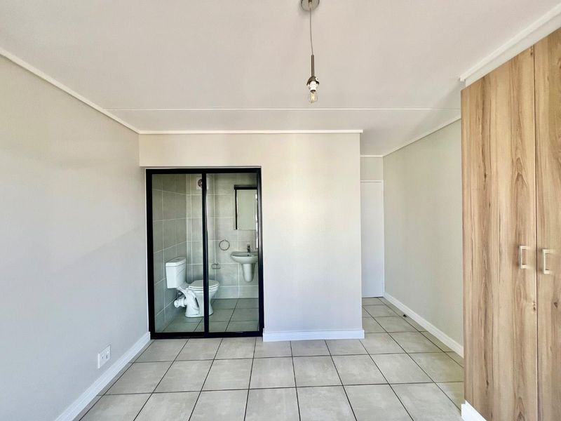 A clean, modern bedroom featuring tiled floors, a built-in wardrobe, and an en-suite bathroom enclosed by a sleek black-framed glass partition.