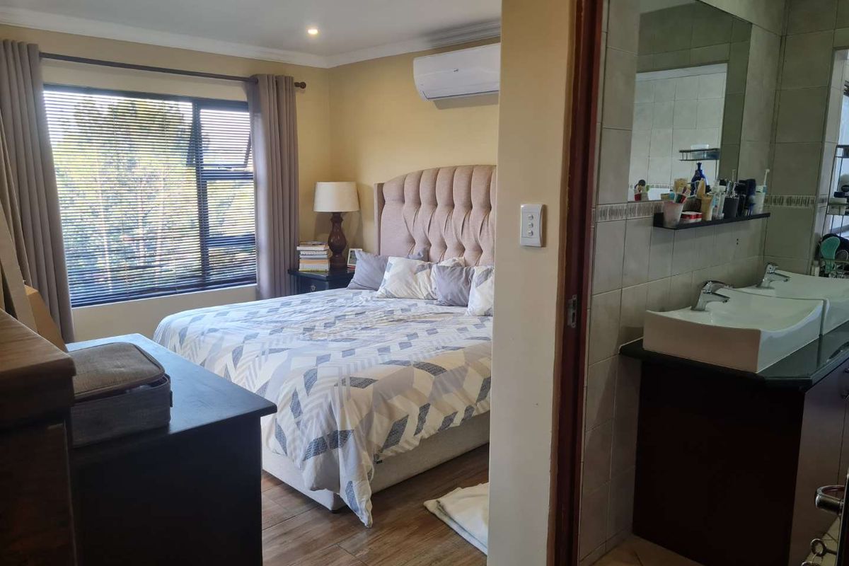 A spacious bedroom featuring wood-look flooring, an integrated air conditioning unit, and an en-suite bathroom with a double vanity and tiled finishes.