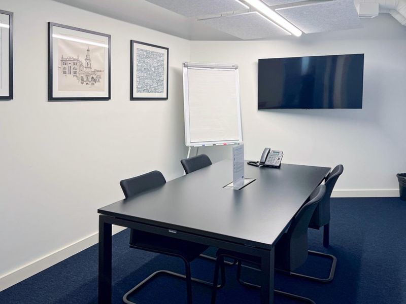 A professional meeting room featuring a dark conference table, wall-mounted display screen, acoustic ceiling tiles, and dark carpeting.