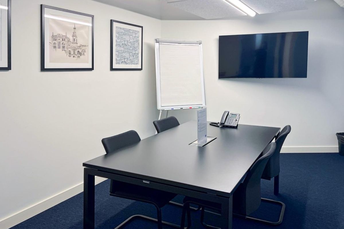 A professional meeting room featuring a dark conference table, wall-mounted display screen, acoustic ceiling tiles, and dark carpeting.
