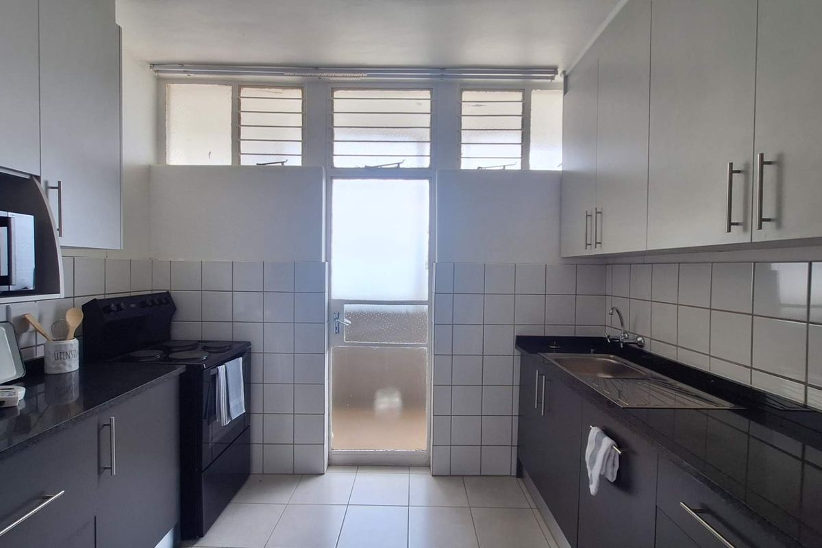 A functional kitchen featuring dark cabinetry, granite countertops, tiled backsplash, and a freestanding stove, with natural light provided by high-level louvre windows.