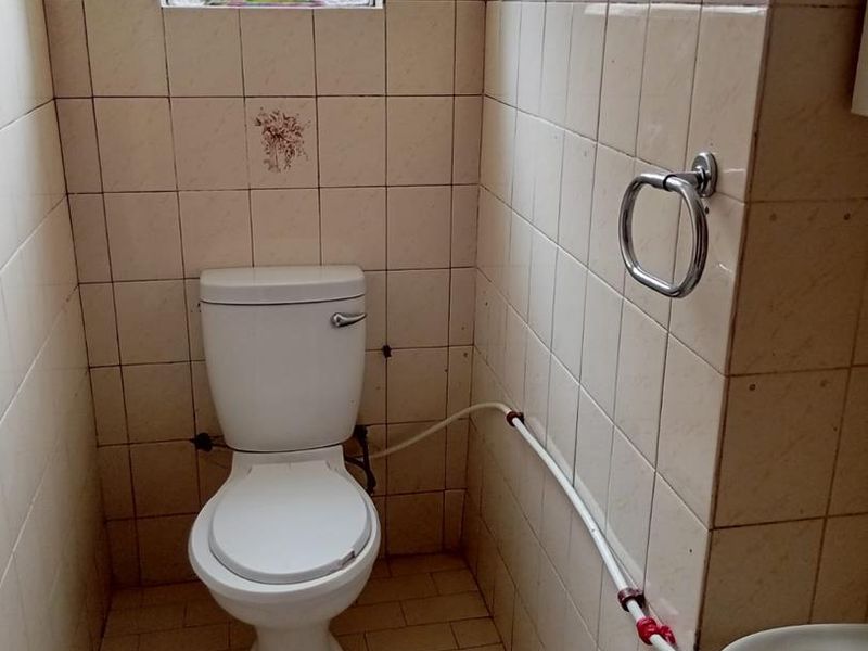 A compact, dated bathroom featuring tiled walls and floor, a standard toilet, and visible surface-mounted plumbing.