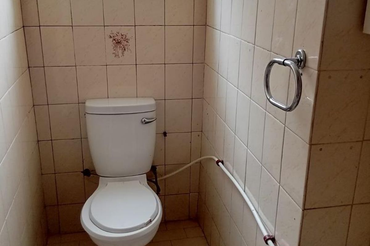A compact, dated bathroom featuring tiled walls and floor, a standard toilet, and visible surface-mounted plumbing.