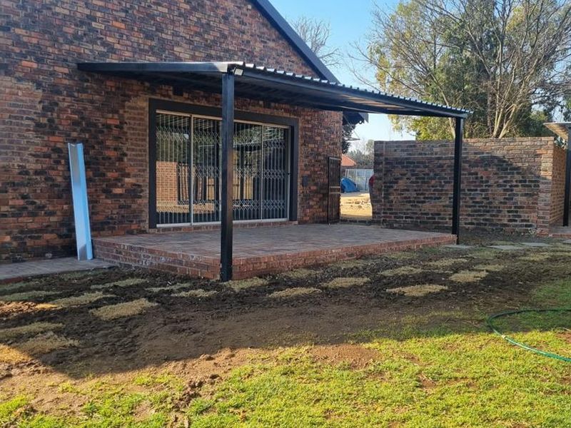 A covered brick patio area featuring a metal roof structure and sliding glass doors, currently adjacent to an unfinished garden space.