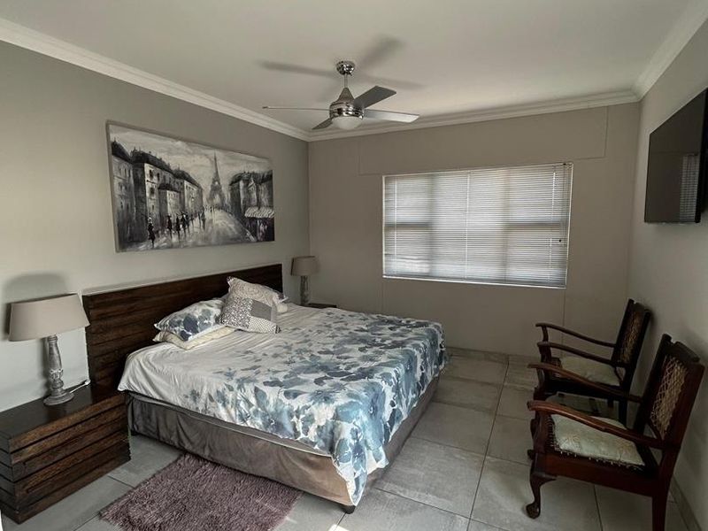 A spacious bedroom featuring neutral tiled flooring, a ceiling fan, and large windows with venetian blinds, offering a clean and well-maintained living space.