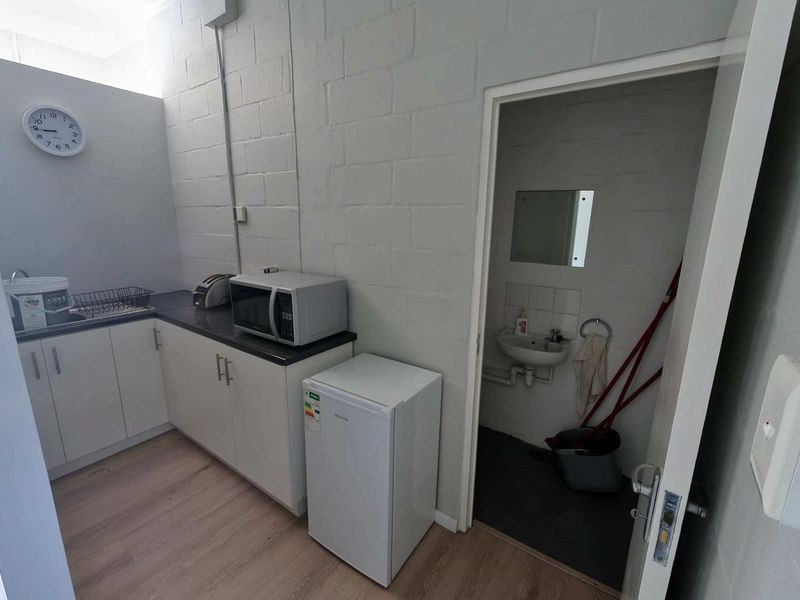 Compact industrial kitchenette featuring white cabinetry, laminate countertops, and an adjoining small washroom with a wall-mounted basin.
