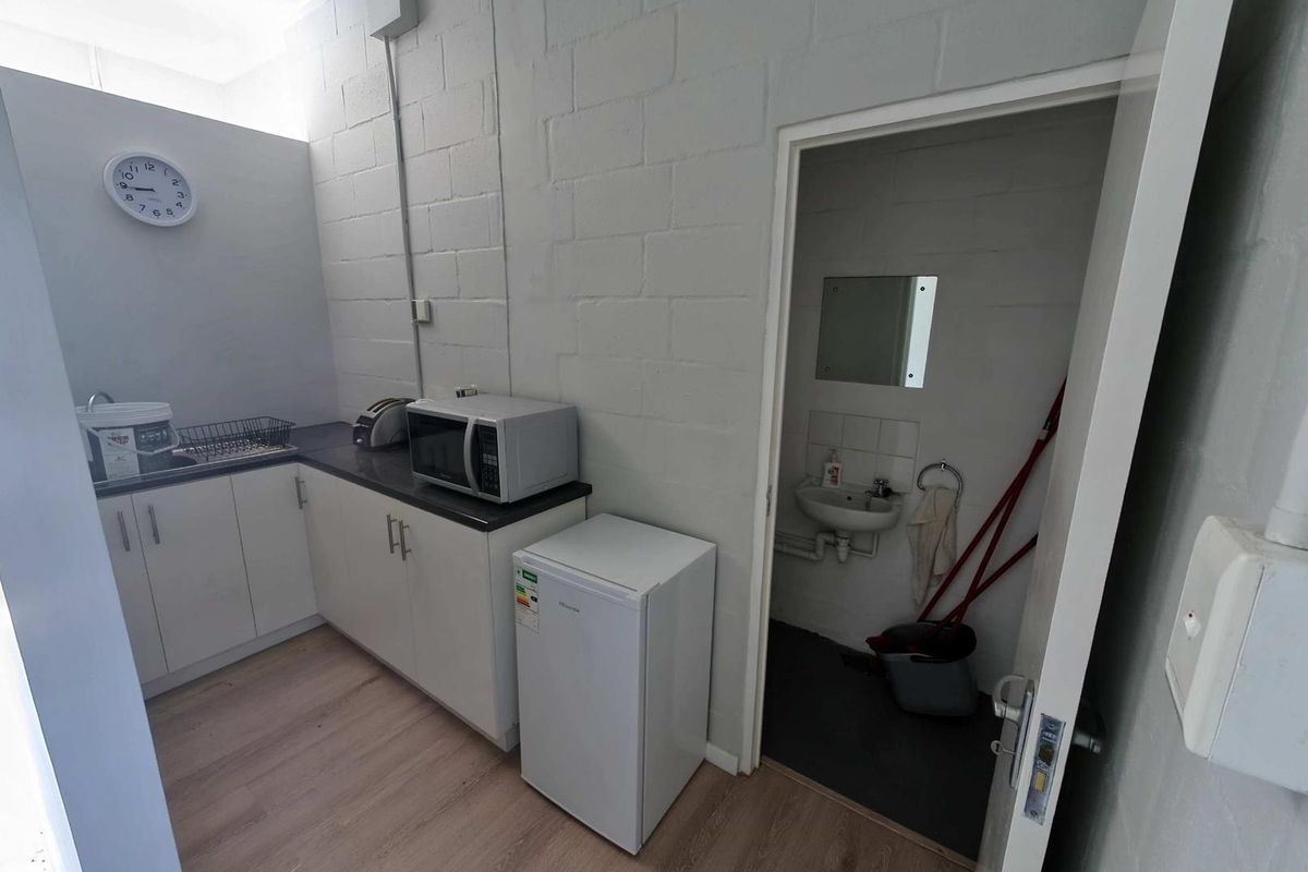 Compact industrial kitchenette featuring white cabinetry, laminate countertops, and an adjoining small washroom with a wall-mounted basin.