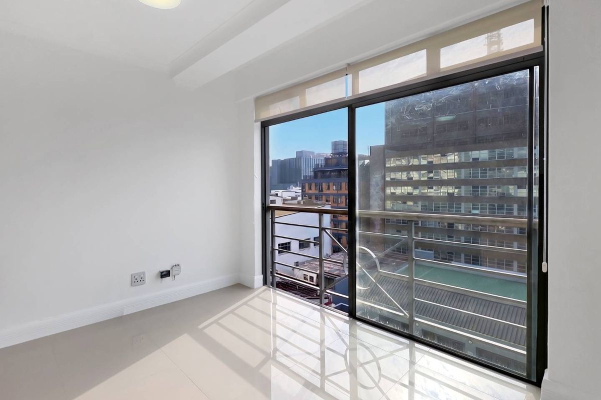 A bright, empty bedroom featuring tiled floors, a ceiling light, and a large sliding glass door opening onto a balcony with urban views.