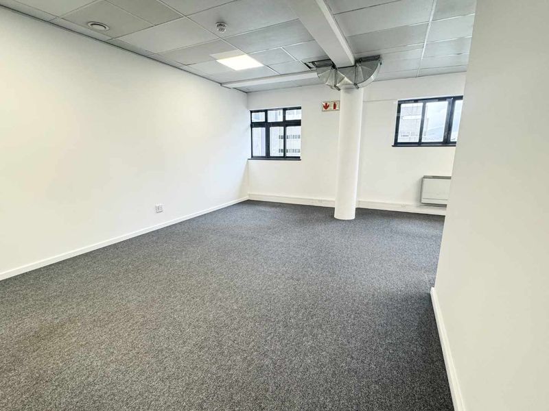 A bright, open-plan office space featuring neutral walls, grey carpeting, suspended ceilings, and large windows providing natural light.