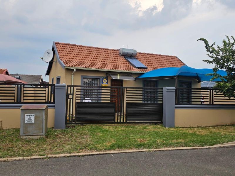 Front view of a single-story house featuring a tiled roof, solar water heater, and a secure perimeter wall with a metal gate.