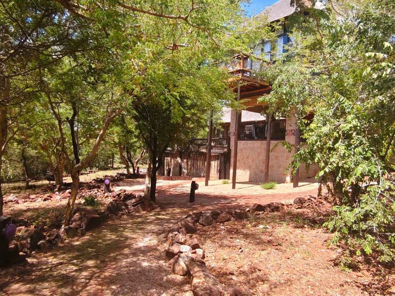 Rear view of the property featuring a raised wooden deck, stone cladding, and a natural garden setting with mature trees and stone pathways.