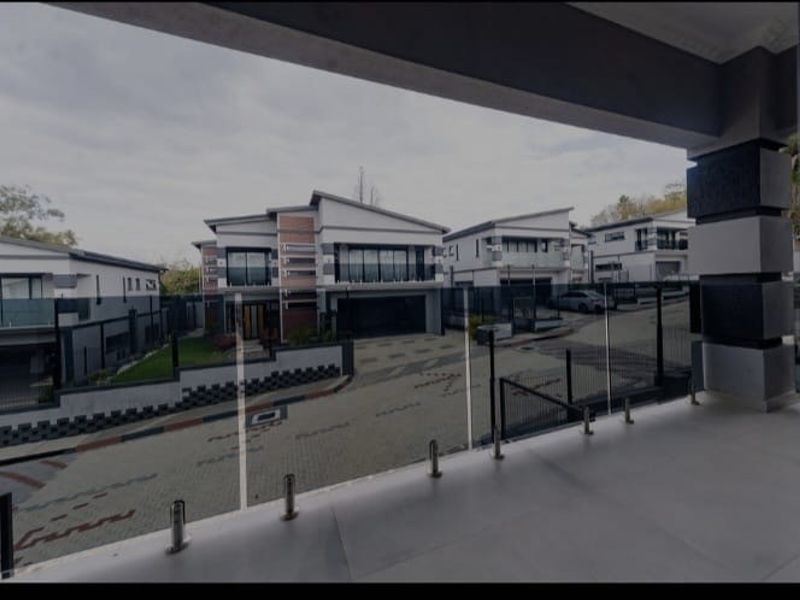 A covered balcony featuring a modern glass balustrade, offering an elevated view of the surrounding townhouse complex and paved driveway area.
