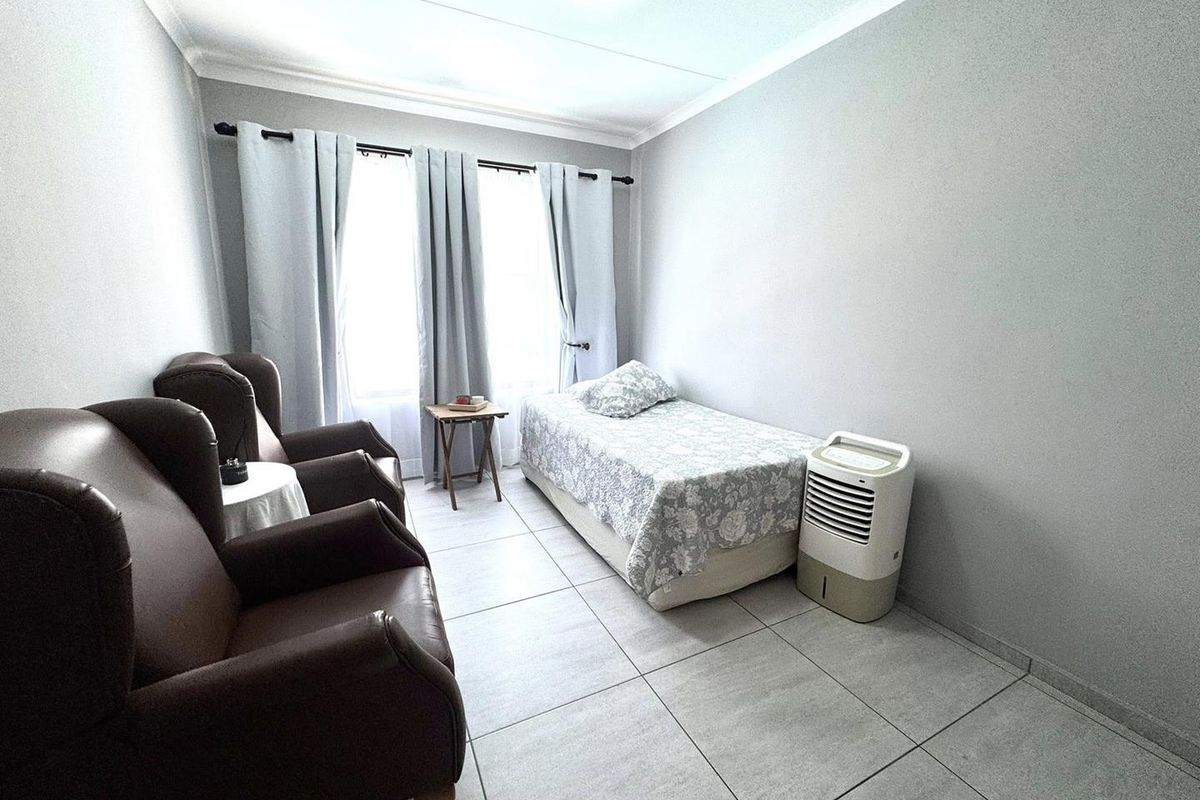 A functional bedroom featuring neutral grey walls, tiled flooring, and large windows with curtain rods, providing a clean and simple living space.