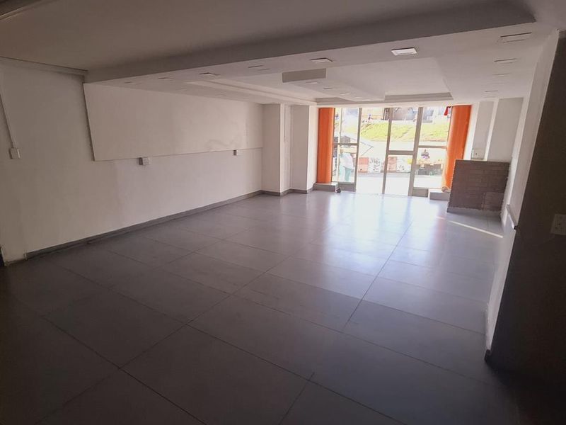 A spacious, empty open-plan commercial or retail space featuring large glass entrance doors, neutral tiled flooring, and recessed ceiling lighting.