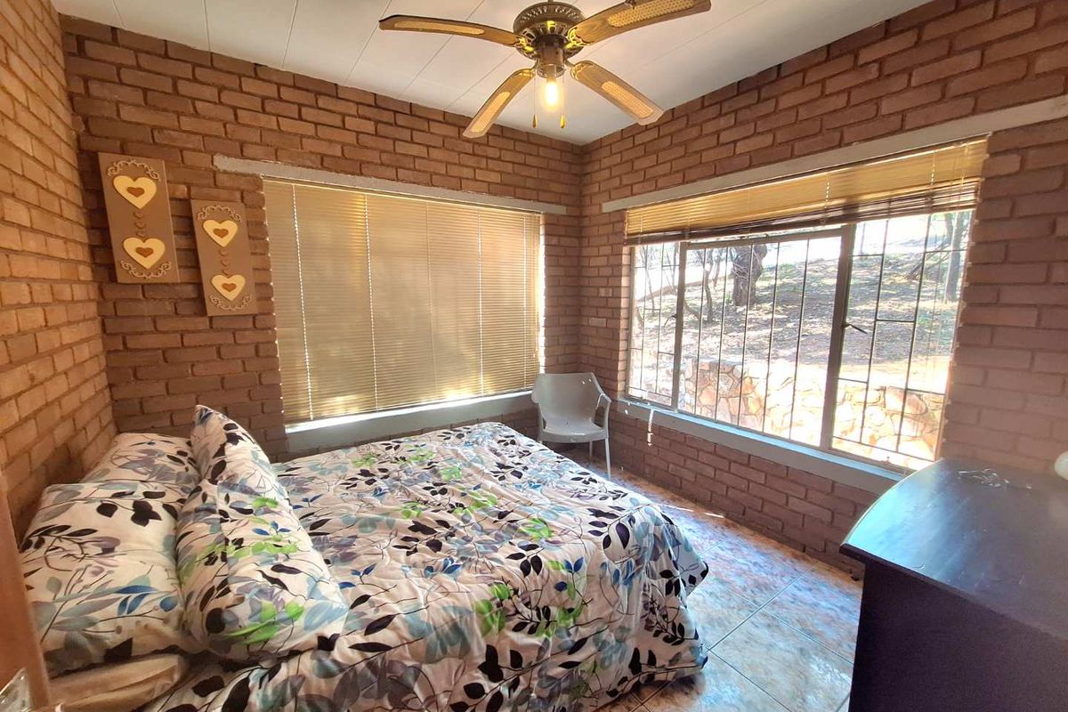 A bedroom featuring exposed brick walls, tiled flooring, a ceiling fan, and large windows with security bars overlooking the garden.