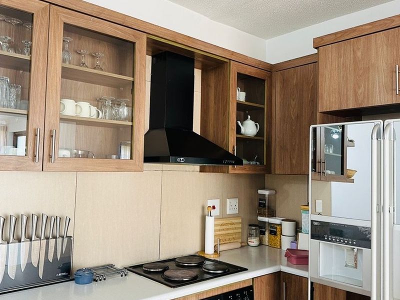 A functional kitchen featuring wood-finish cabinetry, glass-fronted display cupboards, a black extractor fan, electric hob, and built-in oven with a clean tiled backsplash.