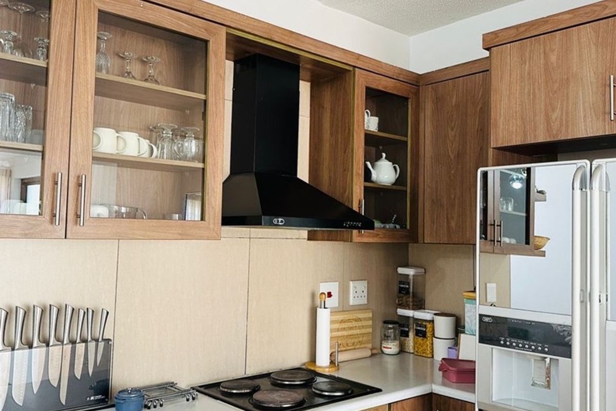 A functional kitchen featuring wood-finish cabinetry, glass-fronted display cupboards, a black extractor fan, electric hob, and built-in oven with a clean tiled backsplash.