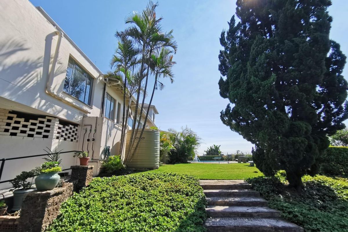 A spacious rear garden featuring a lawn, mature trees, stone steps, and a water tank, with a swimming pool visible in the background.
