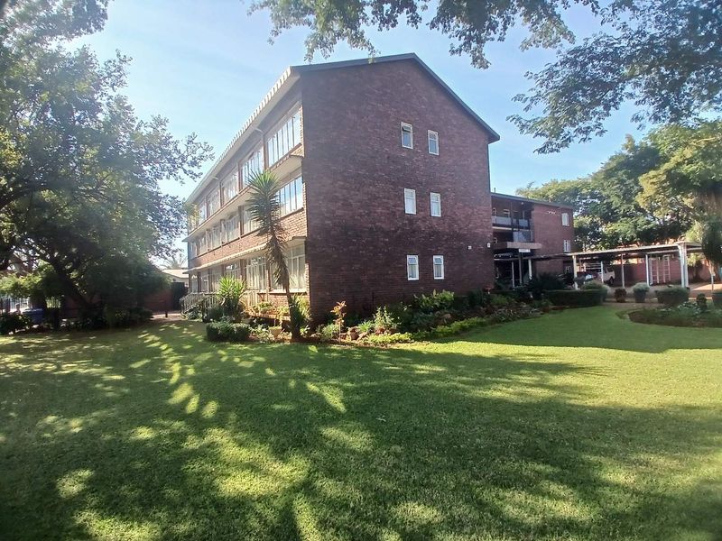 A view of the rear brick apartment building facade, featuring established garden grounds and a covered parking area.