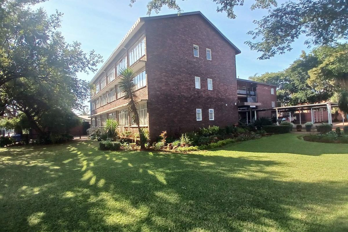 A view of the rear brick apartment building facade, featuring established garden grounds and a covered parking area.