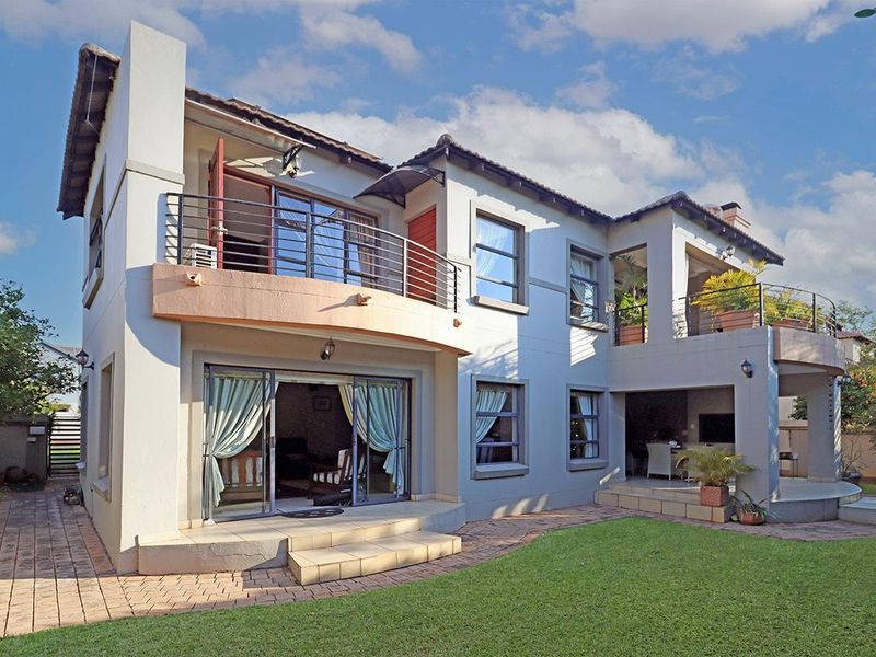 Rear view of a two-story house featuring a covered patio, upper-level balcony with metal railings, and a landscaped garden with paved walkways.