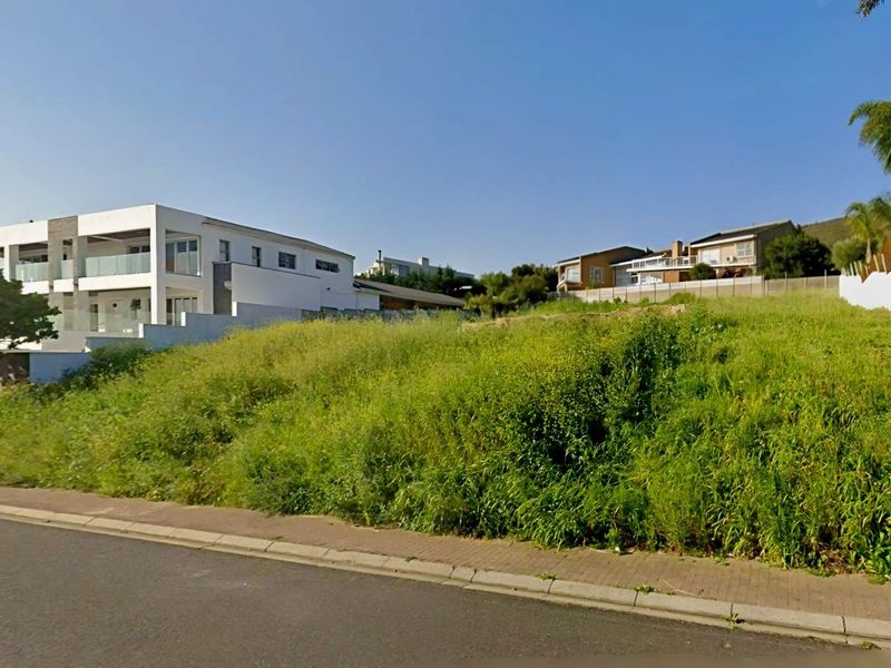 A vacant residential plot featuring a sloped landscape, paved sidewalk frontage, and established boundary fencing in a developed neighborhood setting.