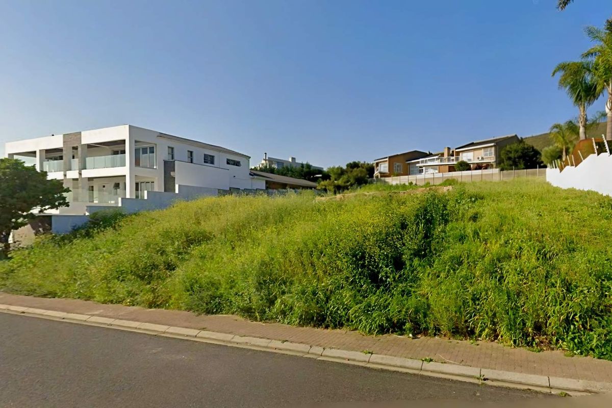 A vacant residential plot featuring a sloped landscape, paved sidewalk frontage, and established boundary fencing in a developed neighborhood setting.