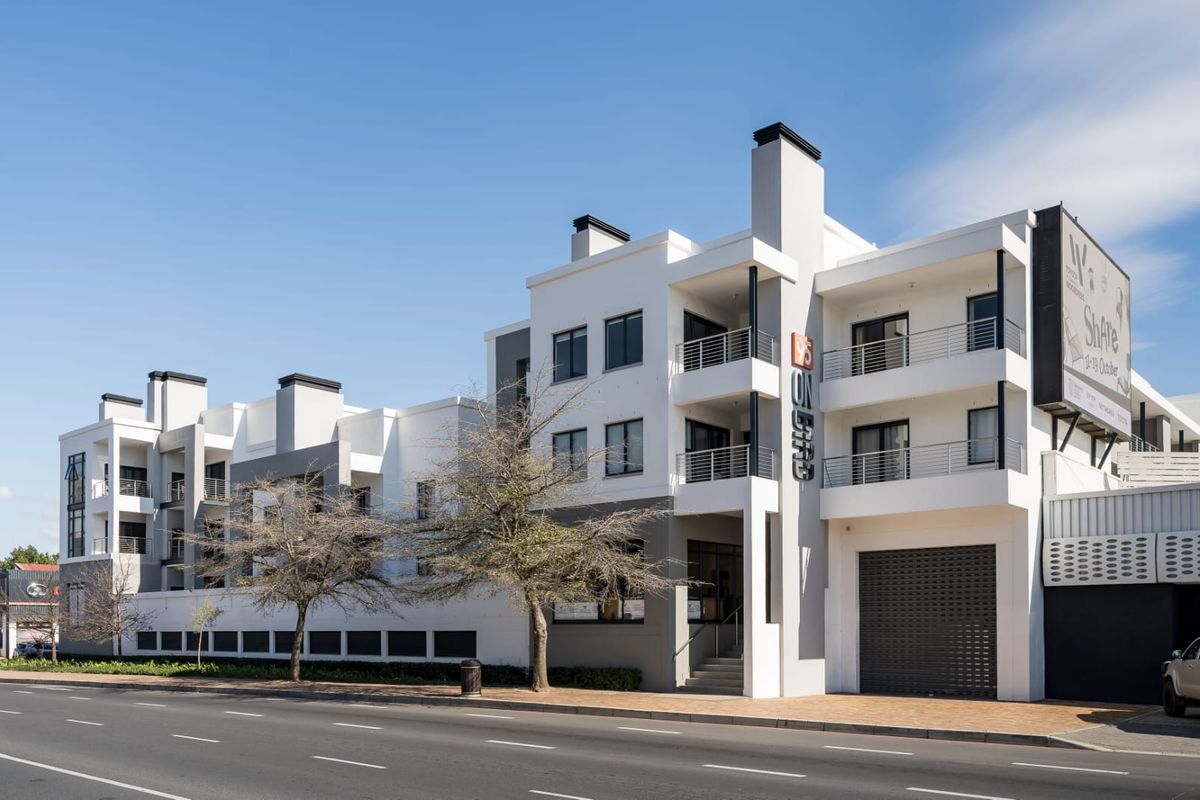 Modern multi-story apartment building exterior featuring clean white and grey facade, private balconies with metal railings, and ground-level roller shutter access.