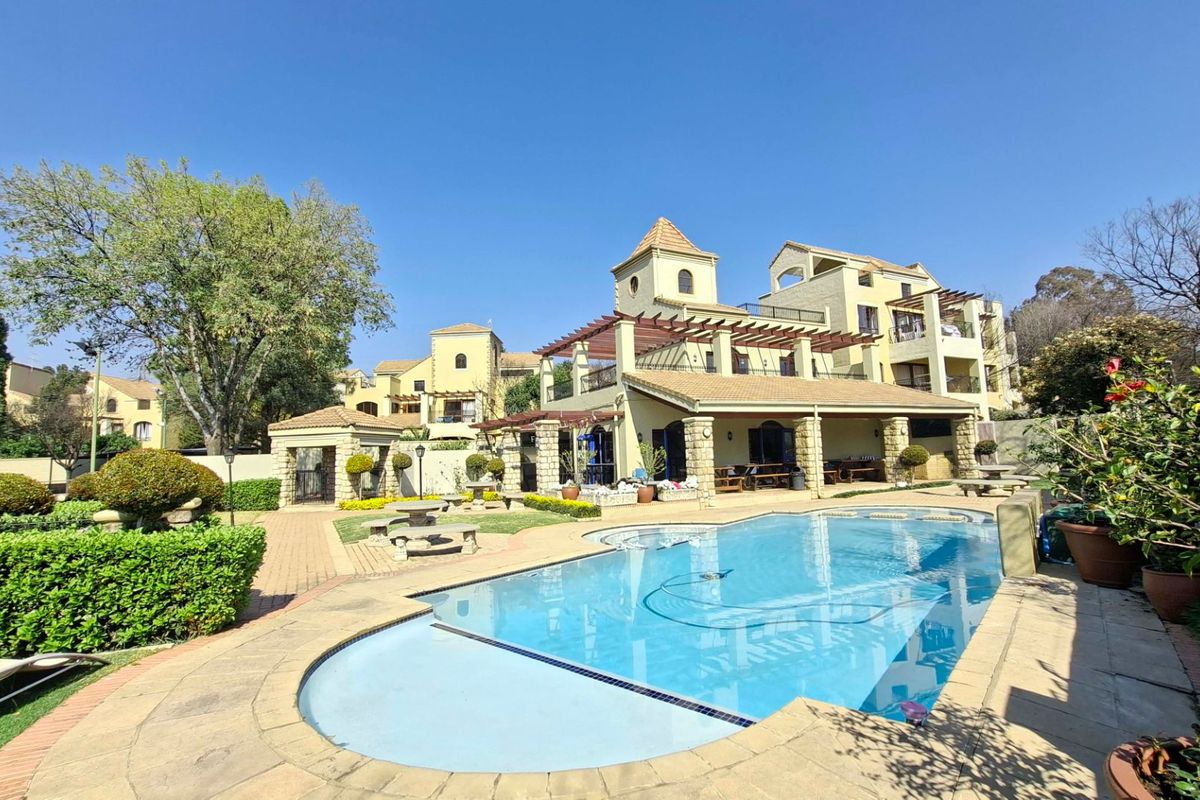 A well-maintained outdoor pool area featuring stone paving, a pergola, and landscaped gardens, set against the backdrop of a multi-story apartment complex.