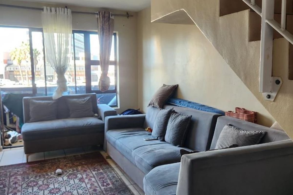 A living room area featuring tiled flooring, a staircase, and large windows providing natural light, currently in need of cosmetic updates.