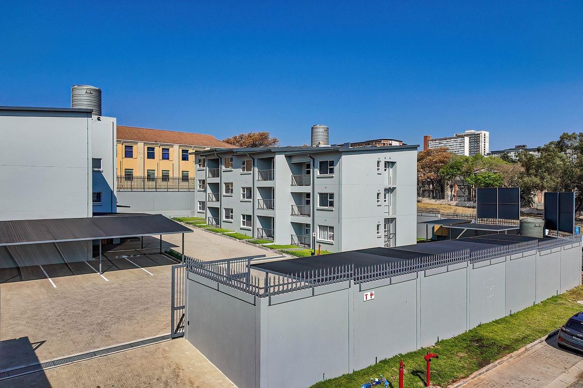 Aerial view of an apartment complex featuring a paved parking area, covered carports, and secure perimeter walling with modern grey exterior finishes.