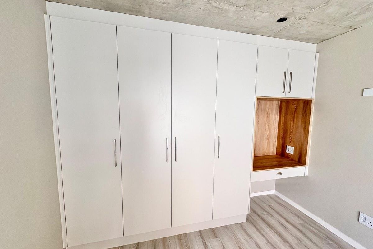 Modern bedroom featuring floor-to-ceiling built-in cupboards with an integrated desk nook, exposed concrete ceiling, and light wood-look laminate flooring.