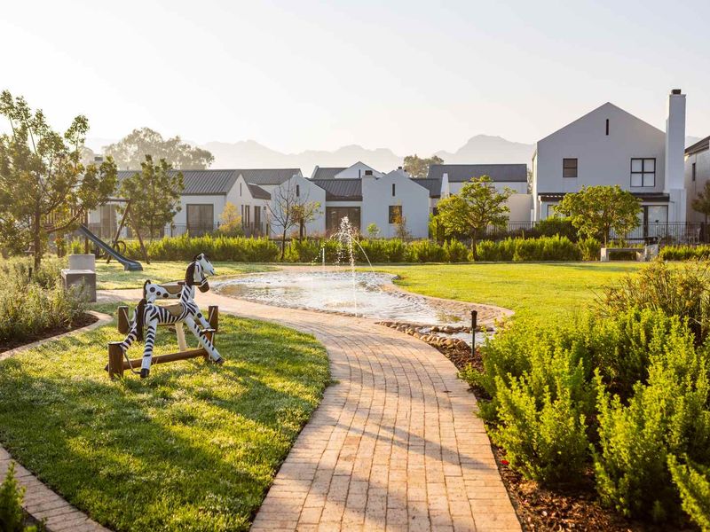 A beautifully landscaped residential garden featuring a brick paved pathway, a central water fountain, and well-maintained lawn areas with surrounding estate homes.