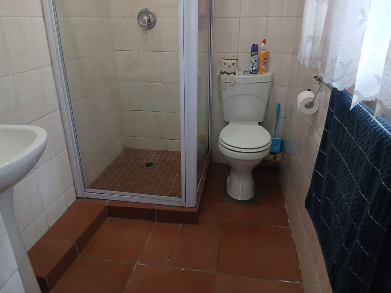 A functional bathroom featuring a glass-enclosed shower, pedestal basin, and toilet with terracotta-style floor tiles and neutral wall tiling.