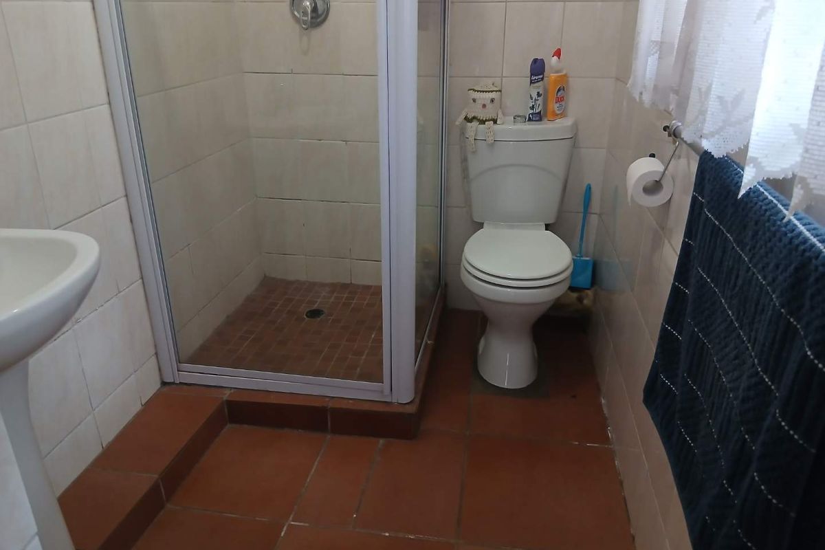 A functional bathroom featuring a glass-enclosed shower, pedestal basin, and toilet with terracotta-style floor tiles and neutral wall tiling.