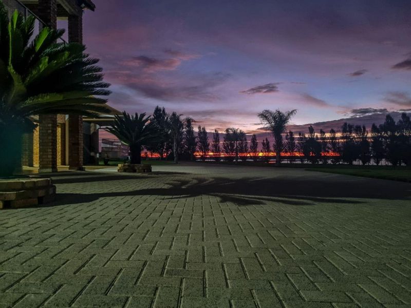 A scenic view of the property's paved driveway and brick facade at sunset, featuring established garden beds and a tranquil atmosphere.