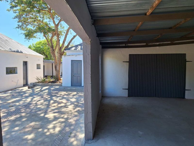 A covered garage area with a concrete floor and corrugated metal door, adjacent to a paved courtyard and guest house buildings.