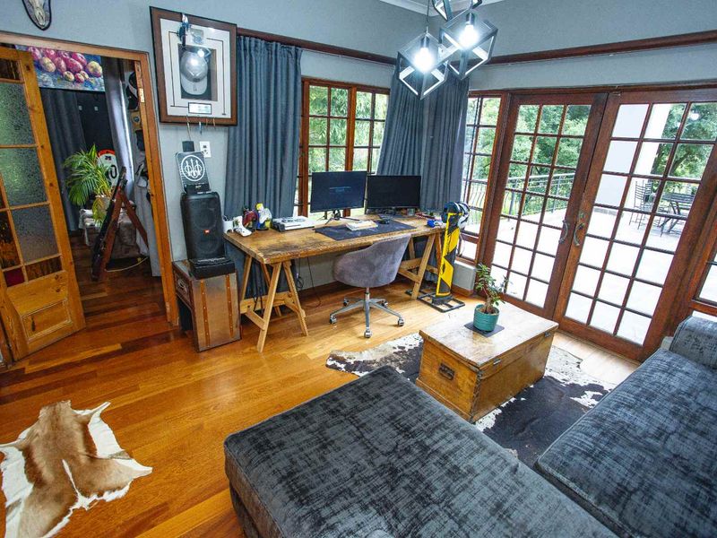 A home office featuring warm hardwood flooring, timber-framed windows, and double french doors leading to an outdoor area.