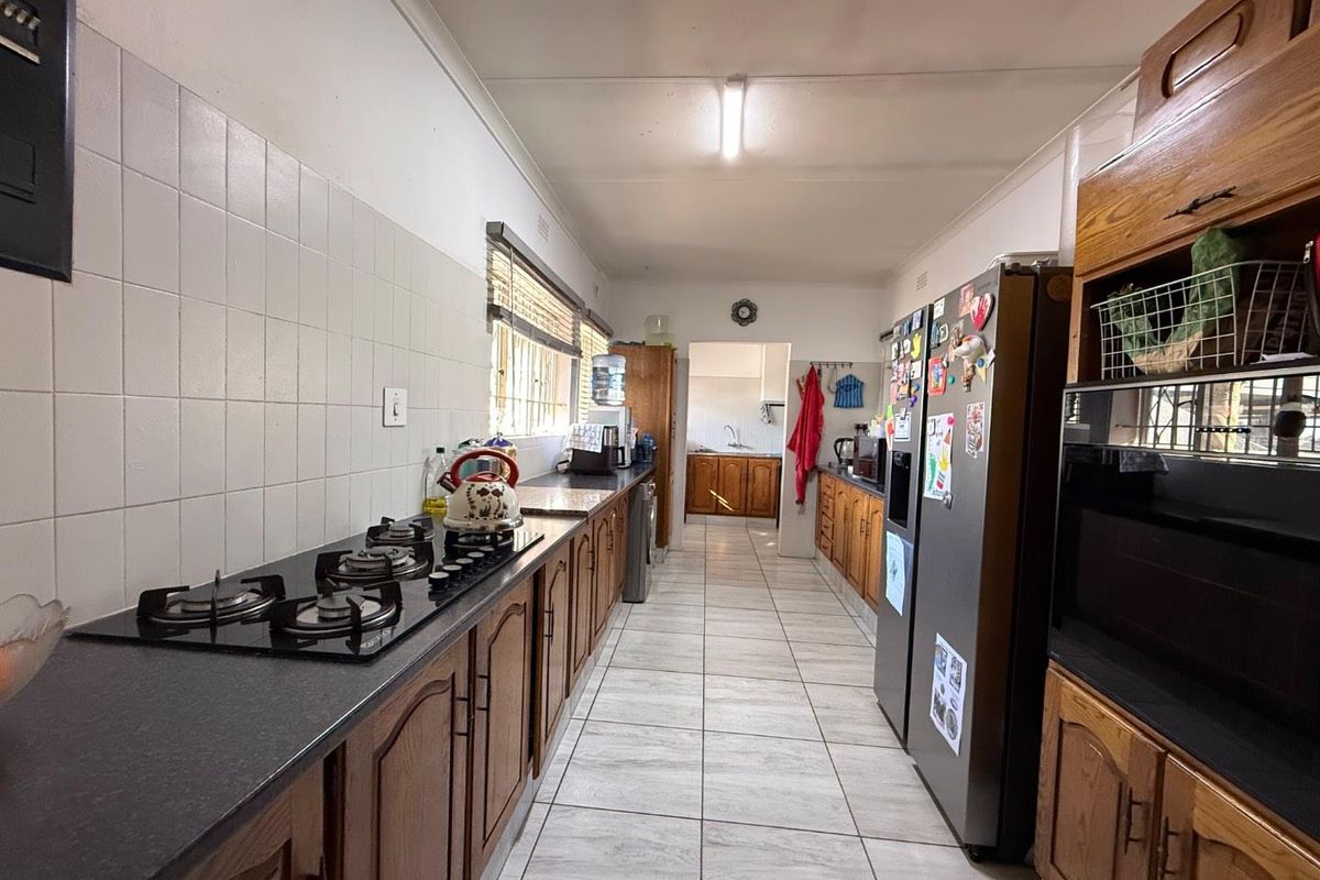 A functional galley-style kitchen featuring wooden cabinetry, a built-in gas hob, tiled flooring, and a tiled splashback, leading into a secondary prep area.