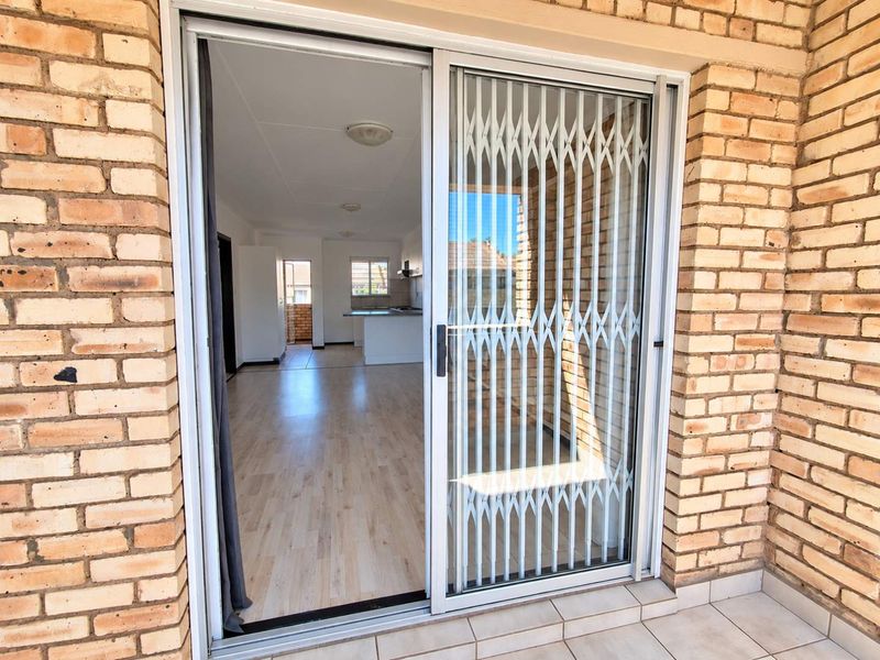 View from a tiled balcony through a sliding glass door with security gate into an open-plan living and kitchen area with laminate flooring.