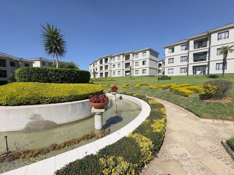 A well-maintained apartment complex featuring landscaped gardens, a decorative water feature, and paved walkways between the residential buildings.