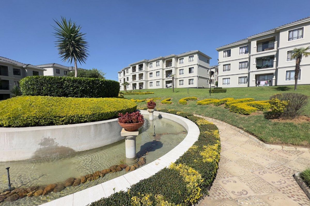 A well-maintained apartment complex featuring landscaped gardens, a decorative water feature, and paved walkways between the residential buildings.