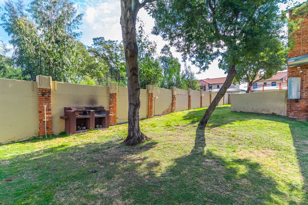 A private rear garden featuring a lawn, mature trees, a built-in brick braai, and a secure perimeter wall with electric fencing.
