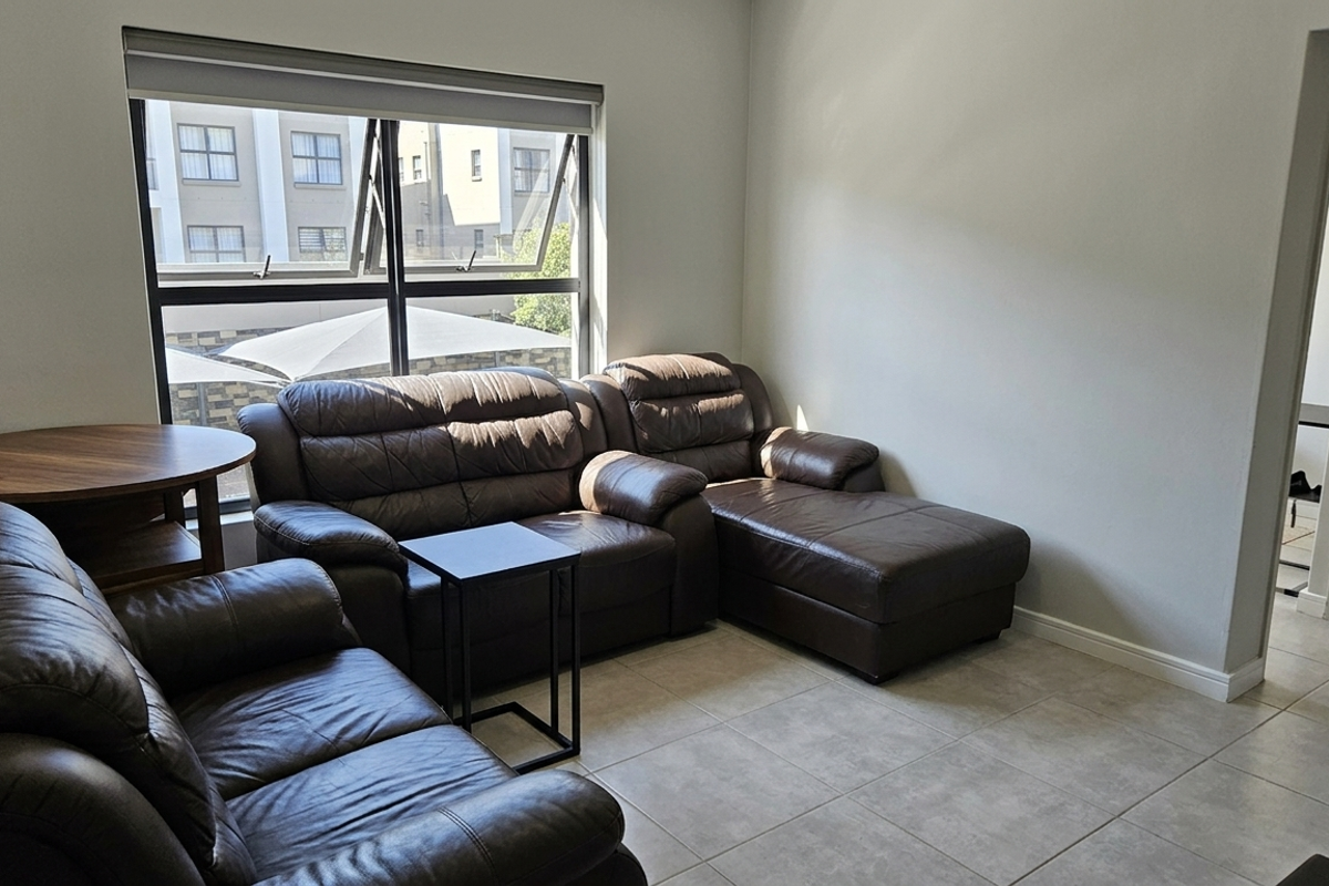 A bright living room featuring neutral tiled flooring, large aluminium-framed windows with roller blinds, and a clean, minimalist aesthetic.