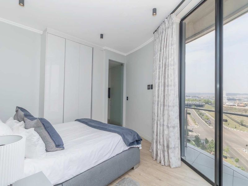 Modern bedroom featuring built-in cupboards, laminate flooring, and a large glass sliding door providing balcony access and expansive city views.