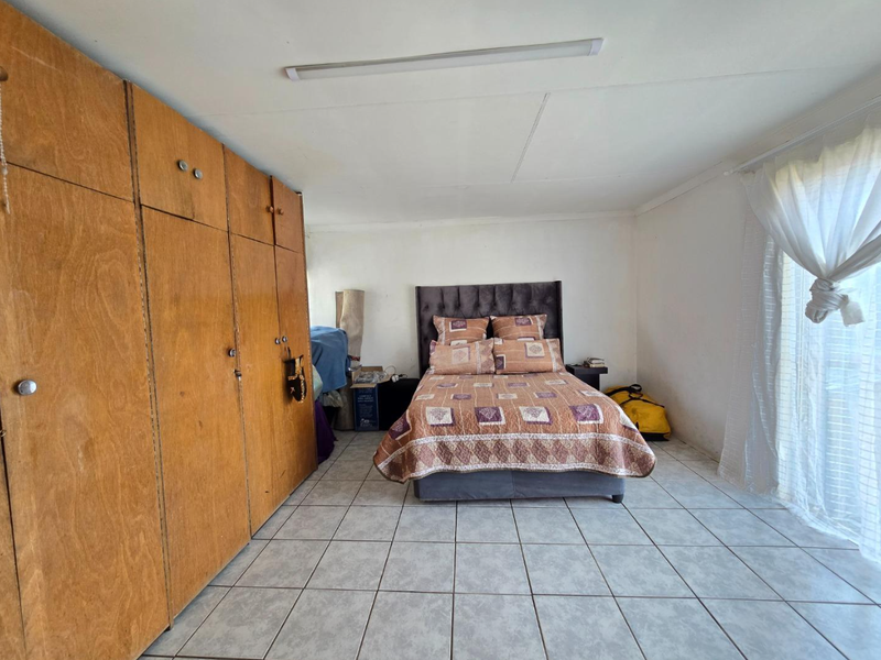 A bedroom featuring built-in wooden cupboards, tiled flooring, and a large window with sheer curtains.