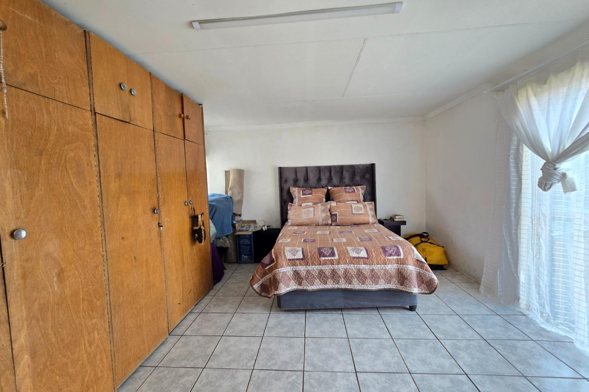 A bedroom featuring built-in wooden cupboards, tiled flooring, and a large window with sheer curtains.