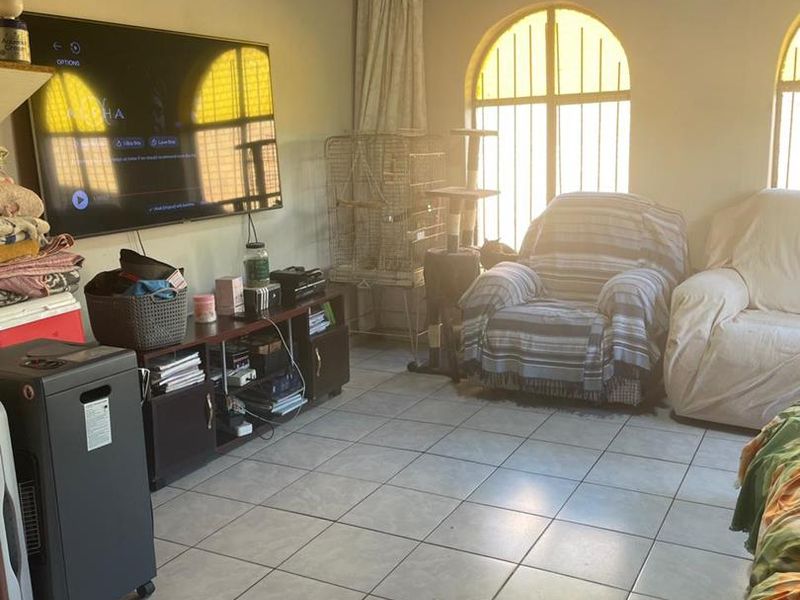 A living room featuring tiled flooring, a timber ceiling, and arched windows with security bars, offering a dated but functional living space.